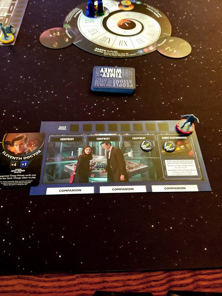 [Image features a game card. The game card shows several spots for dice. Below the dice pool, it shows Clara and The Doctor in the TARDIS. There's a card off to the left where it says "Eleventh Doctor" along with Sonic Screwdriver tokens]